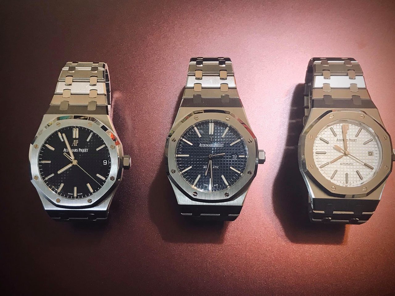 Drei stählerne Audemars Piguet Royal Oak mit achteckiger Lünette und Tapisserie-Zifferblatt in Schwarz, Blau und Silber, liegend vor rotbraunem Hintergrund