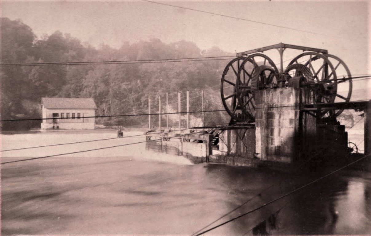 Historische Fotografie des Rheins mit dem Wehr, grossen Seilscheiben der Kraftübertragung und einem Turbinenhaus am Ufer