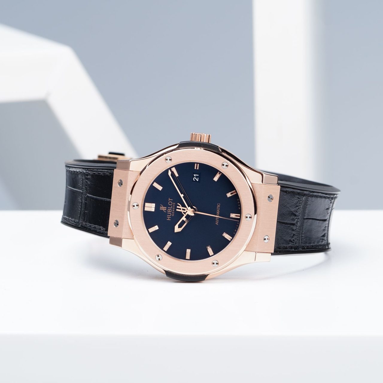 Hublot Classic Fusion mit roségoldenem Gehäuse, sechs H-Schrauben auf der Lünette, dunkelblauem Zifferblatt und schwarzem Band auf weissem Podest