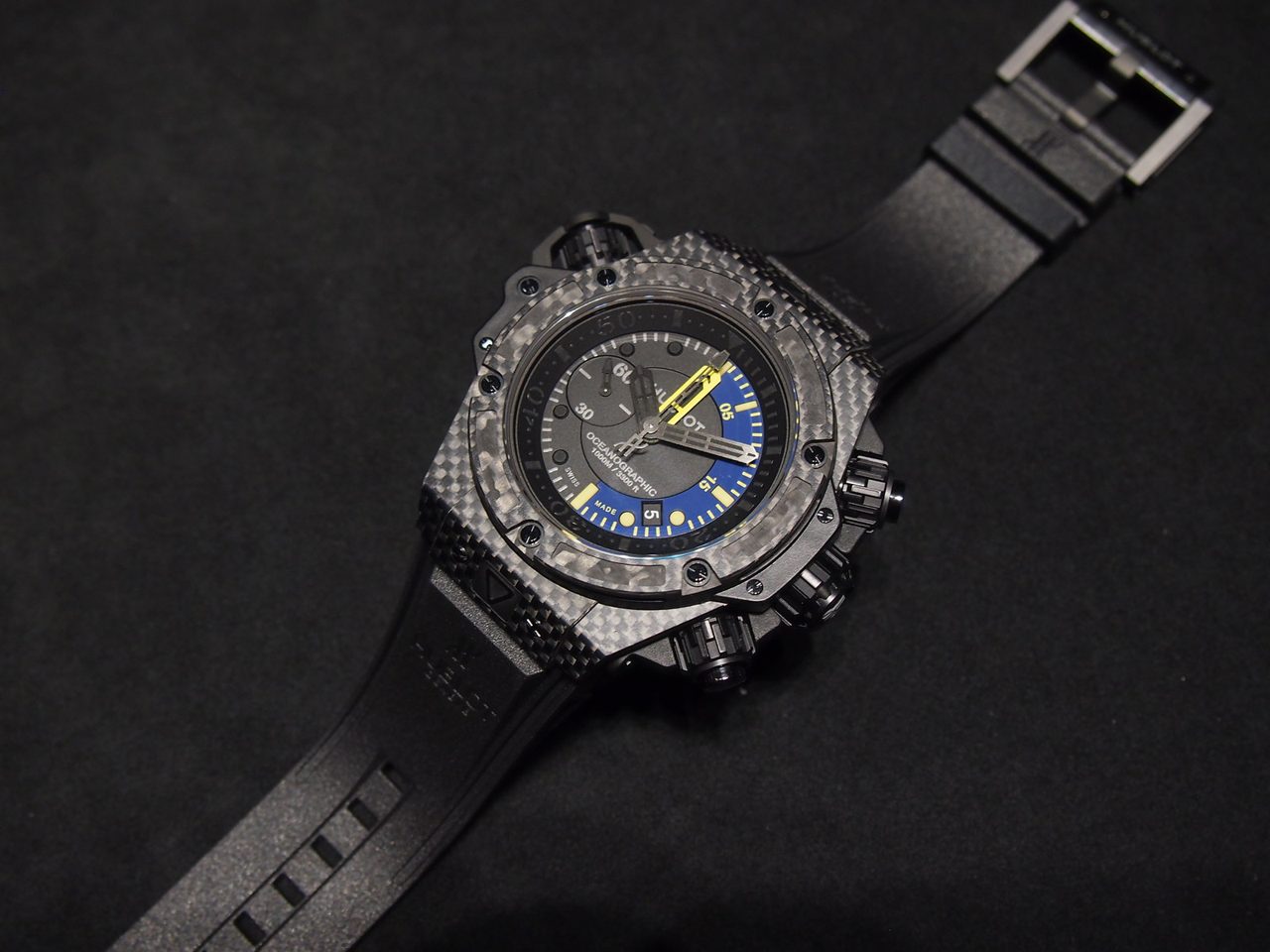 Hublot King Power Oceanographic 1000 mit Carbon-Gehäuse, blau-gelbem Taucher-Zifferblatt und schwarzem Kautschukband, ganze Uhr von oben auf dunklem Untergrund.