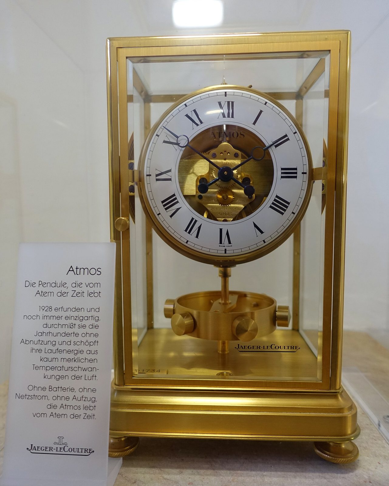 Goldfarbene Atmos-Tischuhr in einem Glasgehäuse mit weissem Zifferblatt, römischen Ziffern und sichtbarem Uhrwerk, daneben eine Informationstafel