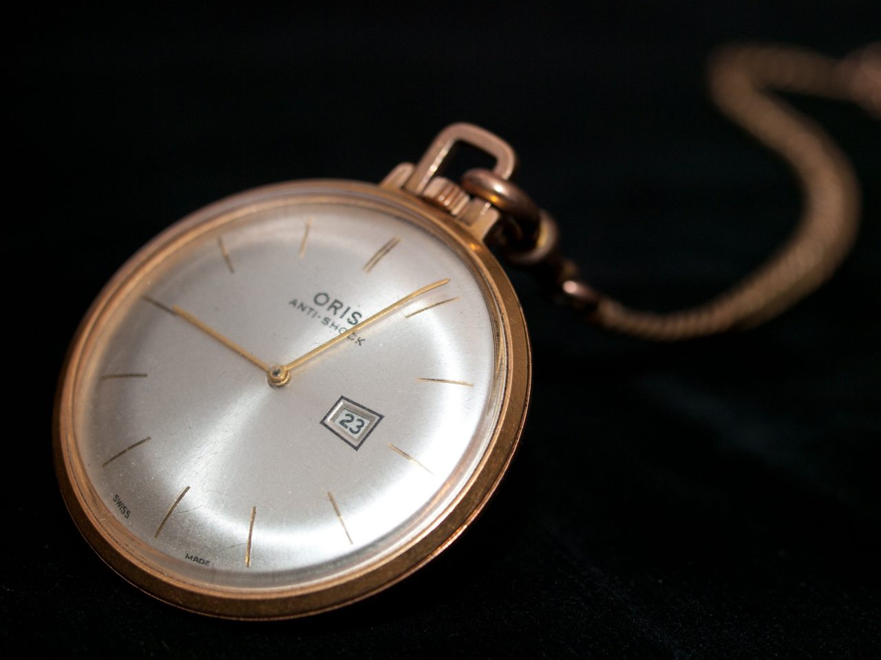Goldfarbene Oris-Taschenuhr mit silbernem Zifferblatt, schlanken Strichindizes, Datumsfenster bei 4 Uhr und der Aufschrift «Oris Anti-Shock», an einer Kette vor schwarzem Hintergrund