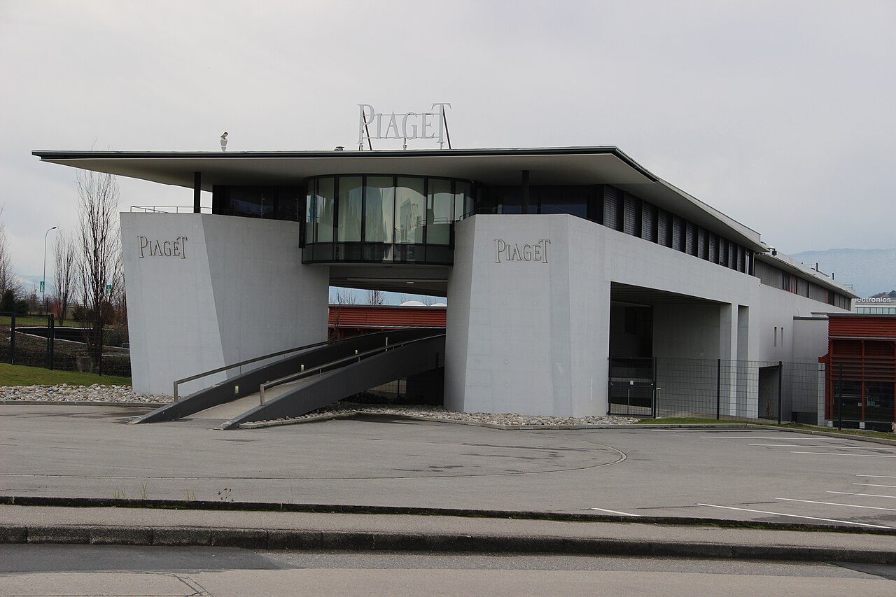 Grauer Betonbau mit auskragendem Dach und dem Schriftzug Piaget auf dem Dach sowie an den Wänden, davor eine Rampe