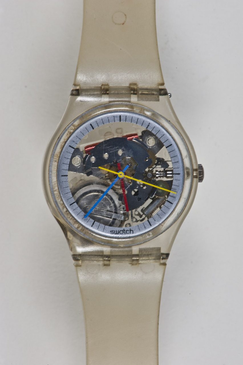 Frühe Swatch mit durchsichtigem Kunststoffgehäuse und hellem Armband; durch das Zifferblatt sind farbige Teile des Werks zu sehen