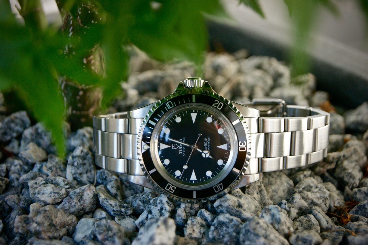 Tudor Oyster Prince Submariner mit Stahlband, liegend auf grauem Granitkies, im Vordergrund unscharfe grüne Blätter.