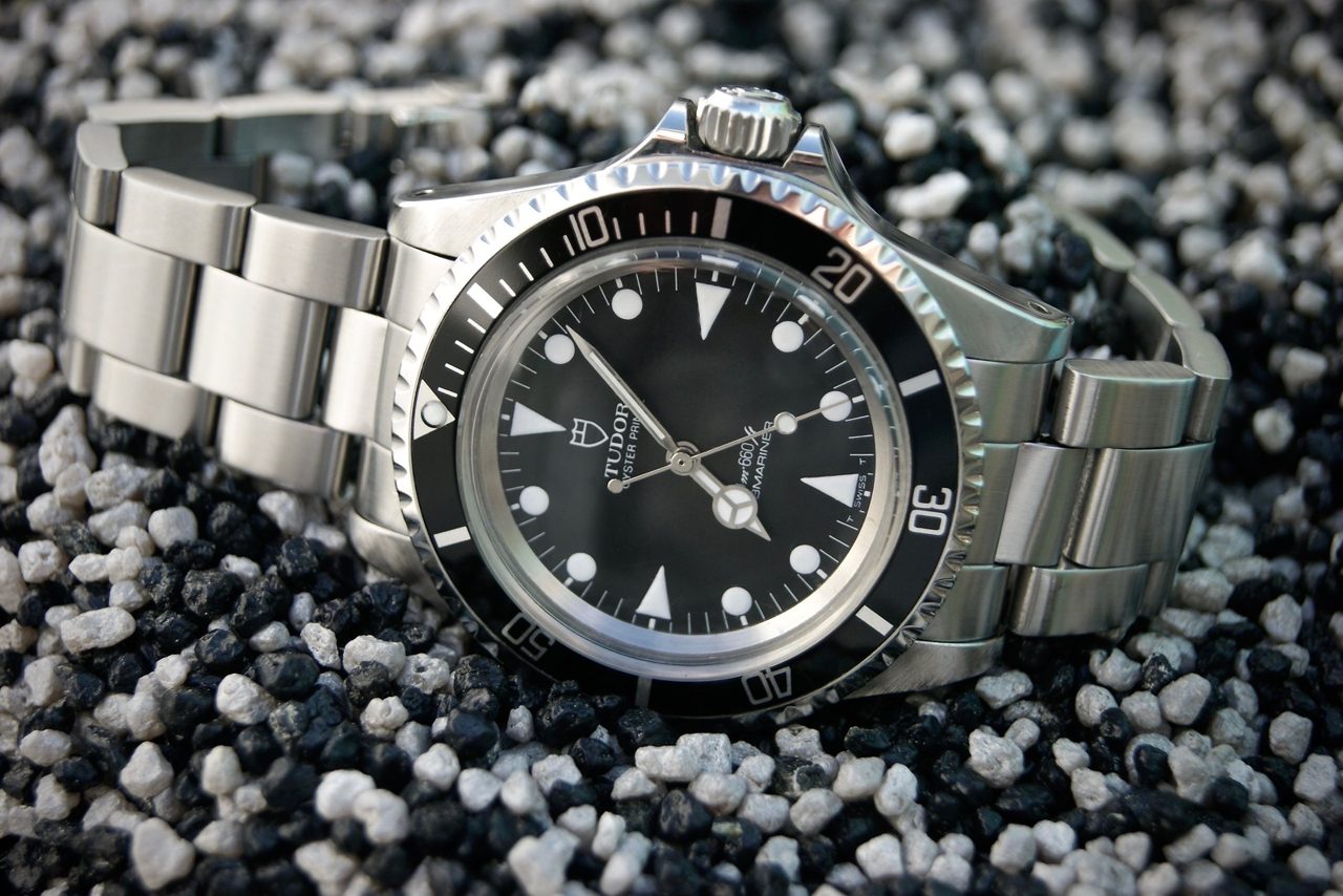Tudor Oyster Prince Submariner mit schwarzem Zifferblatt, schwarzer Lünette und Stahlband, schräg von vorn auf schwarz-weissem Kies fotografiert.
