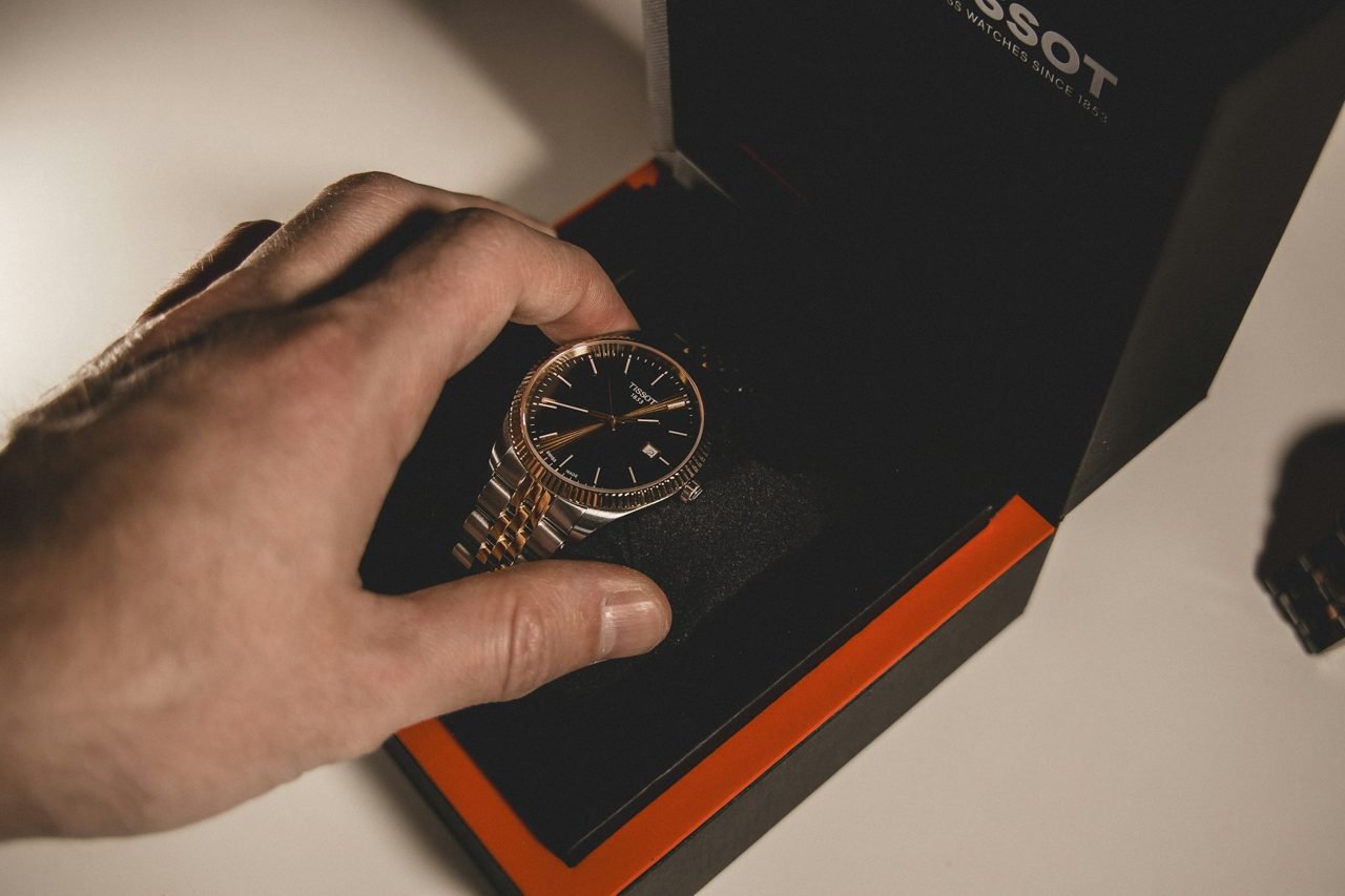 Eine Hand legt eine zweifarbige Armbanduhr in eine schwarze Uhrenbox mit orangem Innenleben, im Hintergrund der Deckel