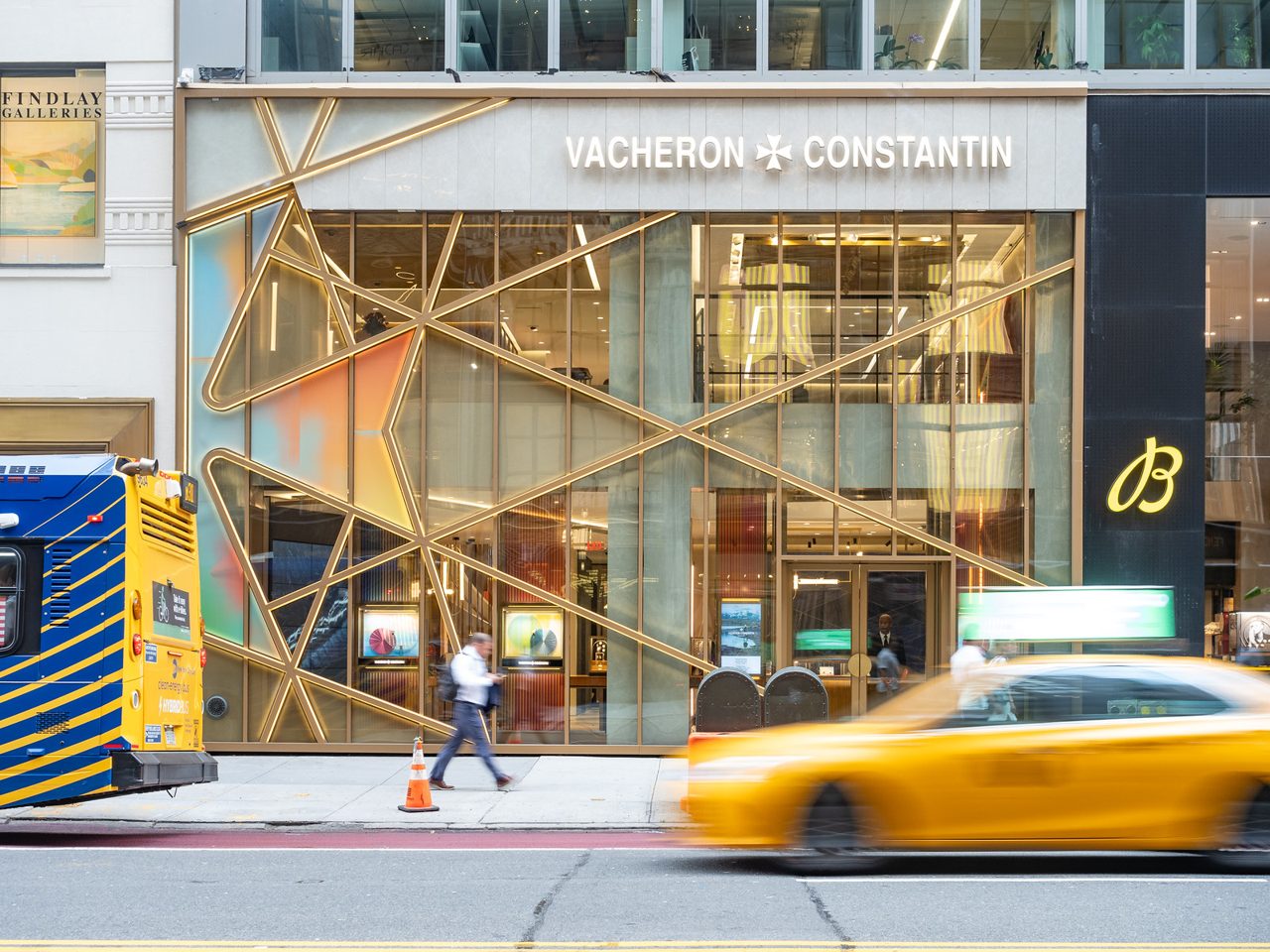 Glasfassade der Vacheron-Constantin-Boutique mit goldenem Malteserkreuz-Gitter und Schriftzug, davor ein vorbeifahrendes gelbes Taxi